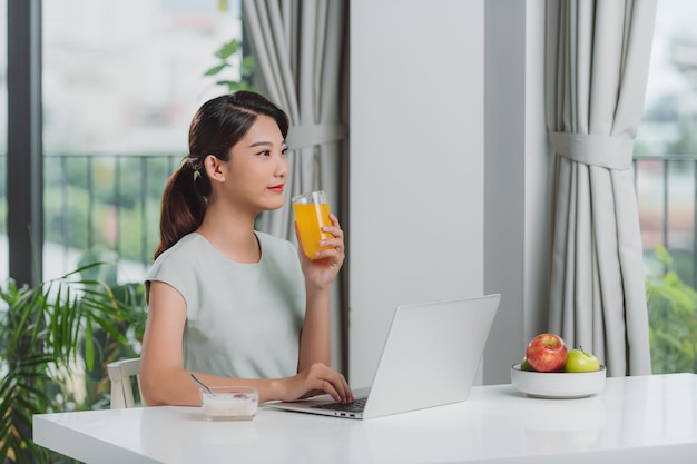 Beautiful young woman working with laptop while breakfast and drinking orange juice