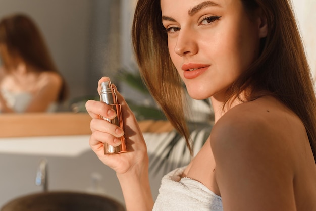 A beautiful young woman, wrapped in a towel, sprays mineral water on her face in front of a mirror. Skin hydration concept.