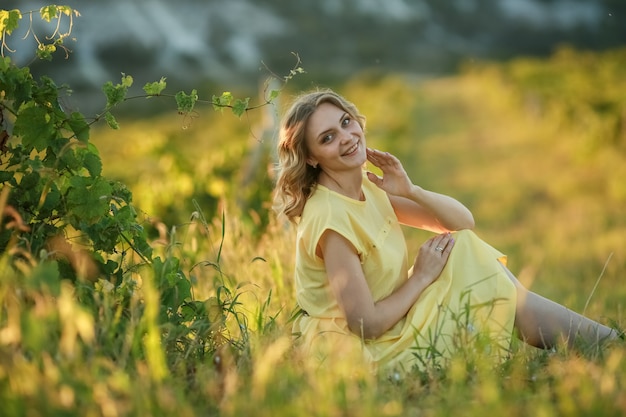 Beautiful young woman in yellow dress sitting on green grass in field