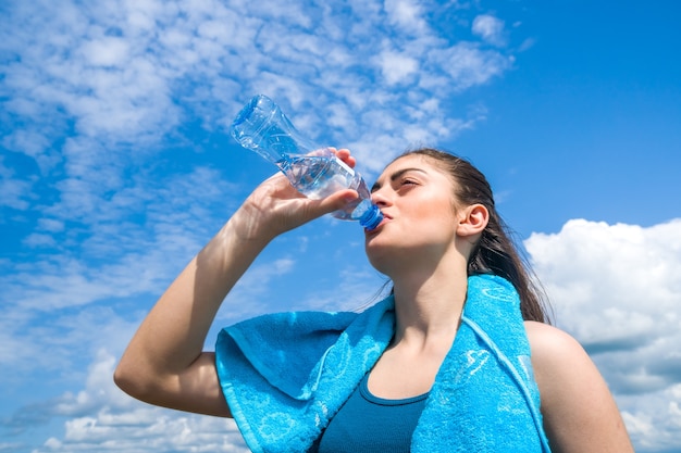 Beautifull girl runner is having break, drinking water against clear blue sky.