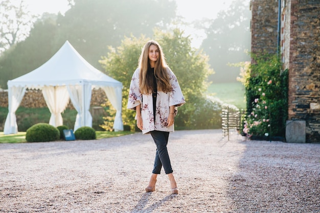 Beautisful stylish woman wears fashionable clothes has long straight hair stand crossed legs against beautiful nature ancient builduing and white tent enjoys sunshine has excursion in unknown place