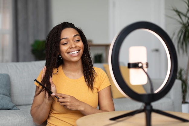 Beauty blog concept African american woman combing her hair and recording video on cellphone talking about hair care