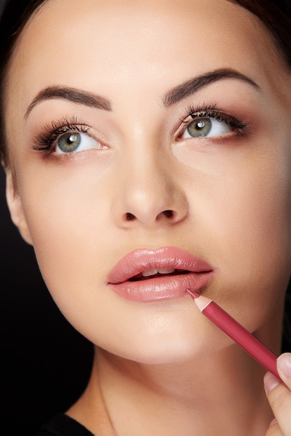 Beauty concept, closeup portrait of woman putting on makeup, painting lips with red lipliner. Portrait of woman touching lips with pencil and looking up, studio, black background