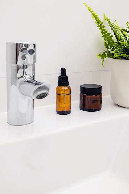 Beauty cosmetic product bottle in brown glass and green potted plant on a white sink in bathroom with copy space Eco friendly sustainable skin care Minimal home interior Morning beauty routine