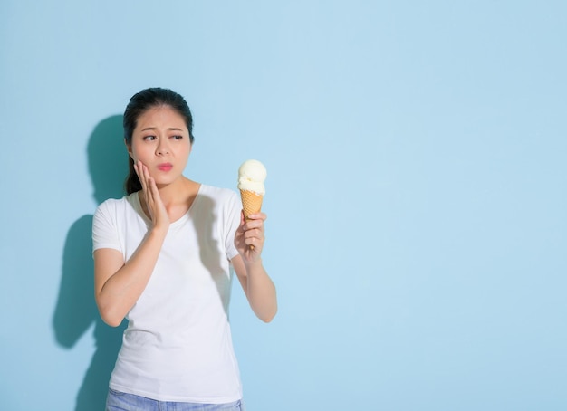 beauty cute female student suffering sensitive tooth disease looking at ice cream hesitate to eating in blue wall background.