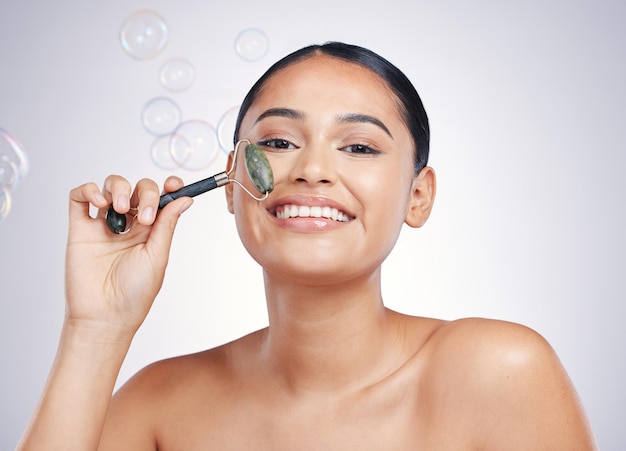 Beauty face roller and a woman in studio for skin glow dermatology or natural cosmetics Portrait of happy female model for facial massage and skincare benefits on bubbles and white background