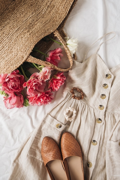 Beauty fashion composition with female sun dress sarafan, shoes, pink peony flowers in straw bag on linen. Flat lay, top view