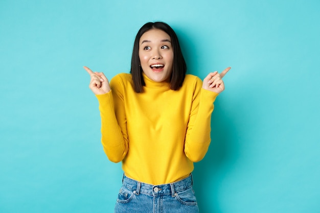 Beauty and fashion concept. Excited asian girl in yellow pullover making choice, pointing fingers sideways and choosing between two options, standing against blue.