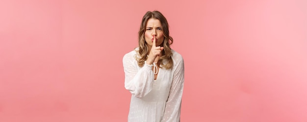 Beauty fashion and women concept Angry seriouslooking blond girl in white dress shushing at someone making noise press finger to lips scolding bad behaviour say be silent pink background