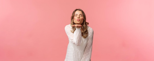 Beauty fashion and women concept Portrait of romantic tender young blond woman in white trendy dress sending air kiss at camera with sassy flirty smile hold hand near lips pink background