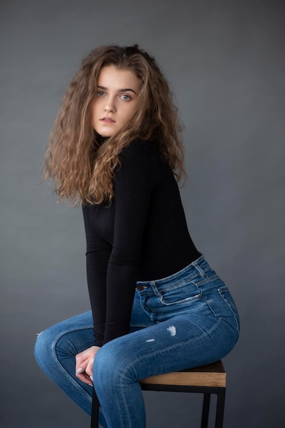 Beauty front image of a young woman with curly hair sitting on high chair on a grey background