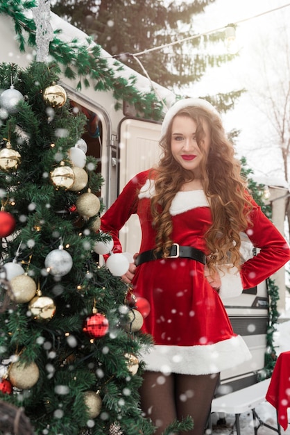 Beauty girl in santa costume decorates the christmas tree at winter campsite getting ready for the n