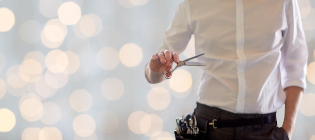 beauty and hair salon, hairstyle and people concept - close up of male stylist with scissors over blank holidays lights background