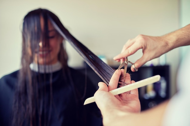 beauty, hairstyle and people concept - happy young woman and hairdresser hands with scissors and comb cutting hair tips at salon