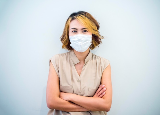 Photo beauty happy asian woman portrait with stylish short hairstyle wearing a white protective face mask and a beige sleeveless shirt stand with crossed arms and looking at camera on a white background