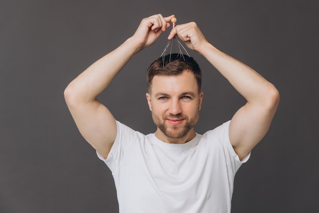 Photo beauty and health hair care a man in a white tshirt does a head massage with a manual massager on a gray background head massage equipment