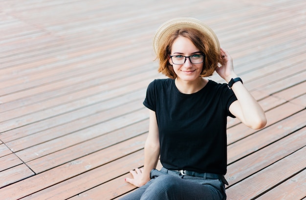 Beauty hipster woman with glasses and hat sitting on wooden floor outdoors