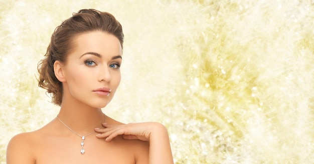 beauty, holidays, people and jewelry concept - woman wearing shiny diamond pendant over yellow lights background