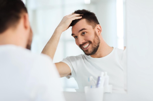 beauty, hygiene, hairstyle and people concept - smiling young man looking to mirror and styling hair at home bathroom