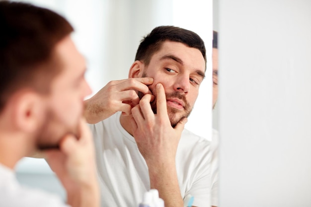 beauty, hygiene, skin problem and people concept - young man looking to mirror and squeezing pimple at home bathroom