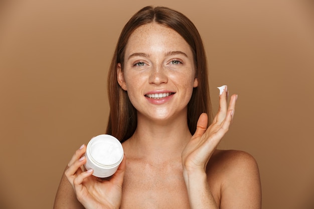 Beauty image of healthy shirtless woman smiling and holding jar with face cream, isolated over beige background