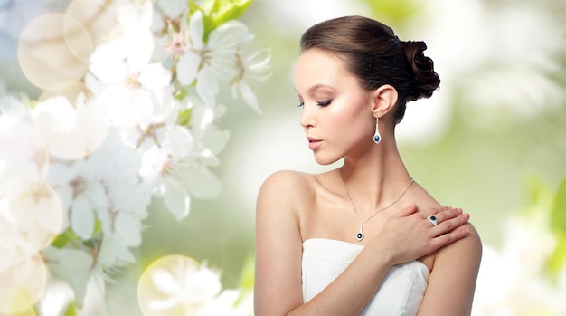 beauty, jewelry, people and luxury concept - beautiful asian woman or bride with earring, finger ring and pendant over natural spring cherry blossom background