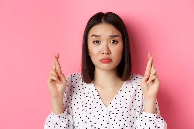 Beauty and lifestyle concept. Close-up of sad and hopeful asian woman making wish, cross fingers good luck and pouting, looking upset, standing over pink background