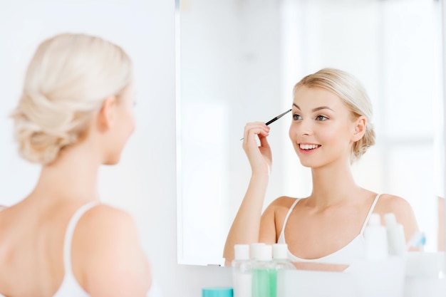 beauty, make up, cosmetics, morning and people concept - young woman applying makeup drawing eyebrow with brush and looking to mirror at home bathroom