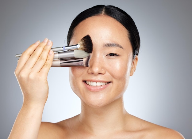 Beauty makeup and woman with brush for cosmetics against a grey mockup studio background Portrait of a young happy and Asian model with a product for facial cosmetic care and mock up space