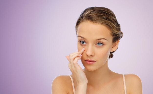 beauty, people, cosmetics, skincare and health concept - young woman applying cream to her face over violet background