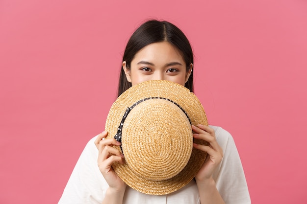 Beauty, people emotions and summer leisure concept. Close-up of shy and cute japanese girl cover face behind straw hat and smiling, standing pink romantic background and blushing coquettish.