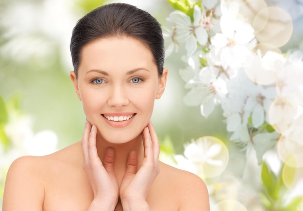 beauty, people and health concept - beautiful young woman touching her face and neck over green blooming garden background