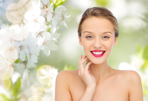 beauty, people and health concept - smiling young woman face with pink lipstick on lips and shoulders over green natural cherry blossom background