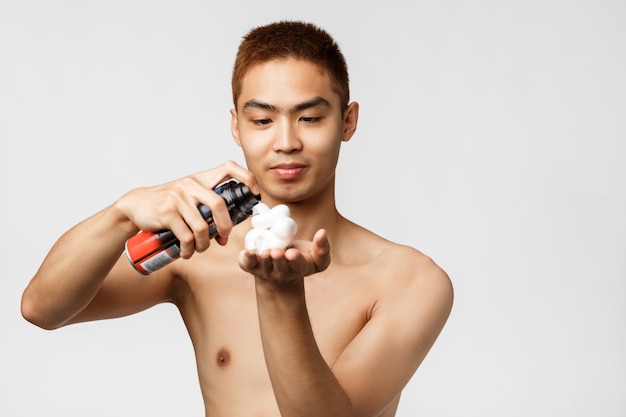 Beauty, people and hygiene concept. Portrait of asian handsome man with naked torso want to shave bristle, smiling pleased as use shaving cream, standing white wall