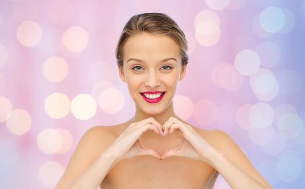 beauty, people, love, valentines day and make up concept - smiling young woman with pink lipstick on lips showing heart shape hand sign over pink lights background