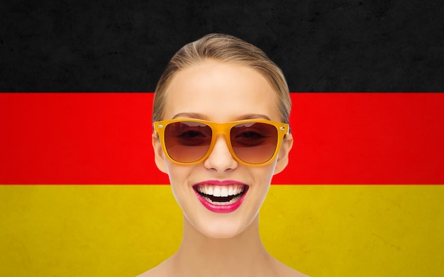 beauty, people, nationality and patriotism concept - smiling young woman in sunglasses with pink lipstick on lips over german flag background