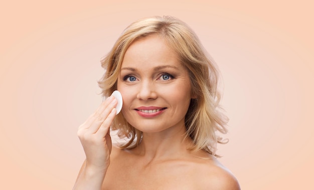 beauty, people and skincare concept - young woman cleaning face and removing make up with cotton pad over beige background