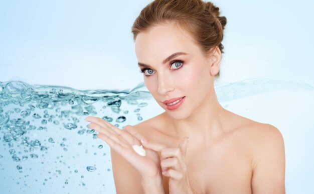 Photo beauty, people, skincare and cosmetics concept - happy young woman with moisturizing cream on hand over blue background with water splash