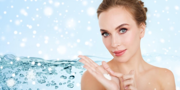 Photo beauty, people, skincare and cosmetics concept - happy young woman with moisturizing cream on hand over water splash bubbles on blue background and snow