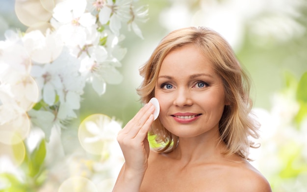 beauty, people, skincare and natural cosmetics concept - young woman cleaning face and removing make up with cotton pad over cherry blossom background