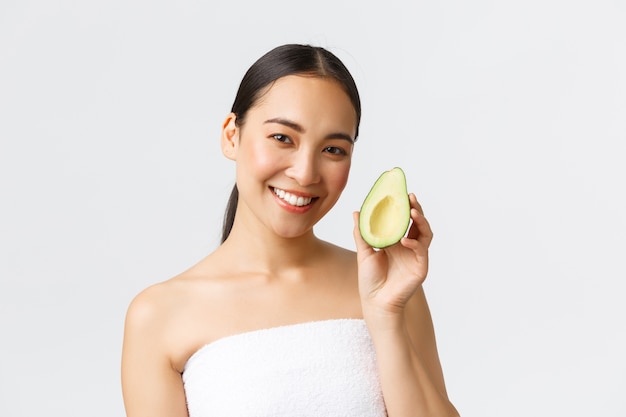 Beauty, personal care, spa and skincare concept. Close-up of beautiful asian female in bath towel showing avocado and smiling, recommend face mask or facial hydrating and nourishing cream.