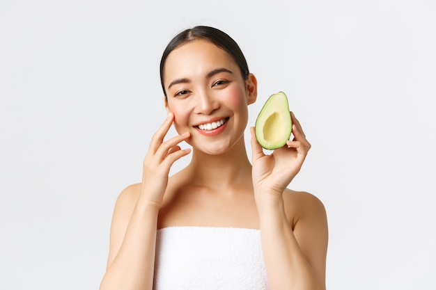 Beauty, personal care, spa and skincare concept. Close-up of beautiful asian woman in bath towel gently touching face and showing avocado as recommend nourishing face mask, cleanser or cream.