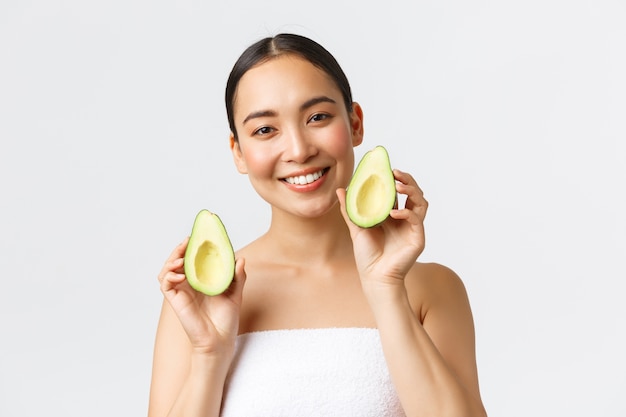 Beauty, personal care, spa and skincare concept. Close-up of tender feminine asian woman in bath towel, smiling and showing avocado near face, advertisement of face mask, cleanser or cream.