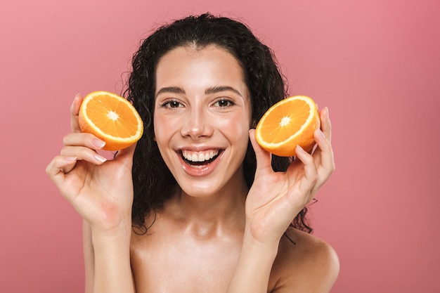 Beauty photo of happy half naked woman with long hair smiling and holding pieces of orange, isolated over pink background