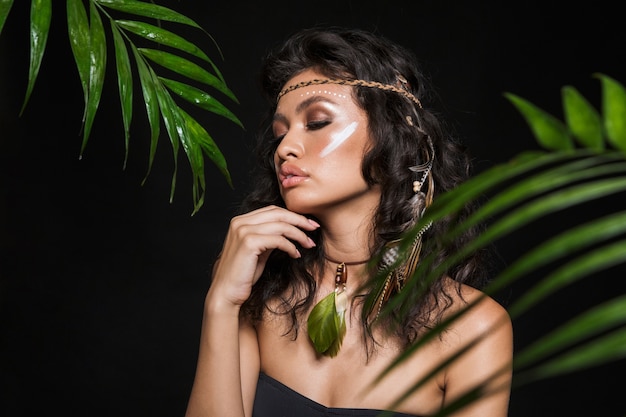 Beauty portrait of an attractive sensual young brunette woman wearing tribal accessories posing with tropical leaves isolated