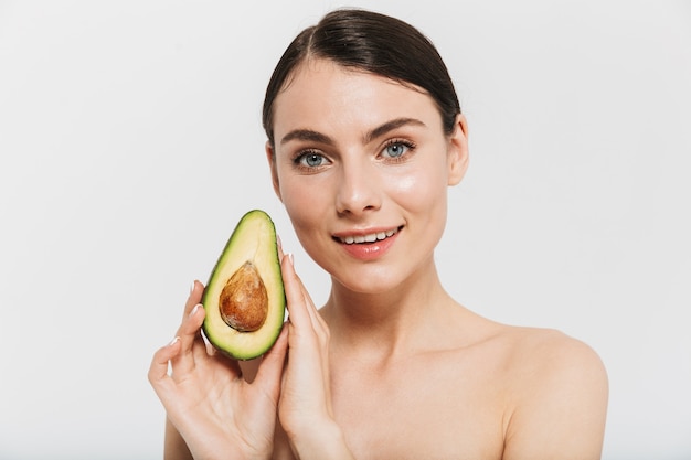 Beauty portrait of an attractive young topless woman isolated over white wall, showing sliced avocado