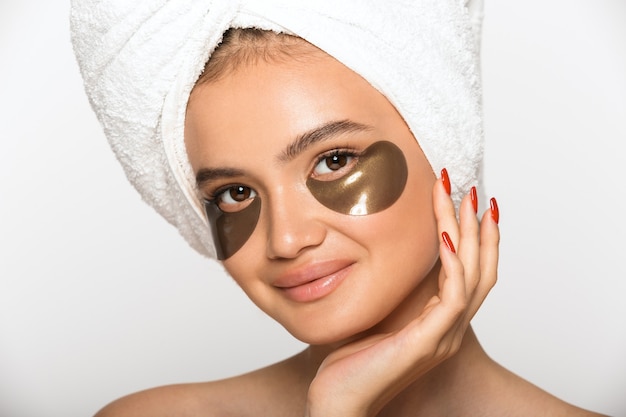 Beauty portrait of an attractive young topless woman wearing bathroom towel on her head standing isolated over white wall, wearing cosmetic eye patches