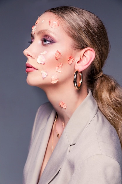 Beauty portrait of a girl Beautiful woman in a jacket posing with flower petals on her face on a gray background Femininity and beauty