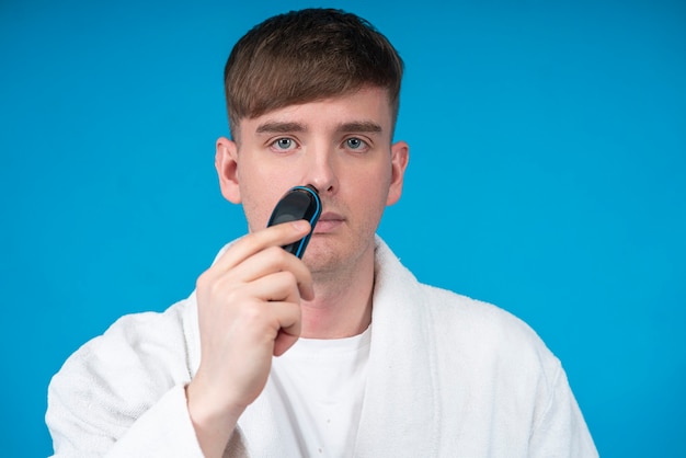 Beauty portrait of handsome guy young man in white bath robe using electric razor or shaver to