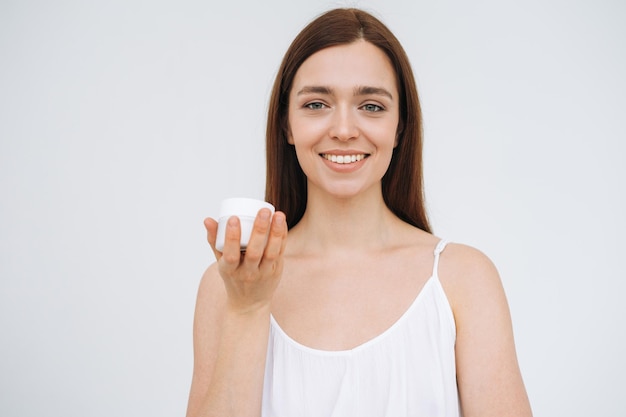 Beauty portrait of happy smiling woman with dark long hair put day nourishing moisturizer cream on clean fresh skin face and hands on the white background isolated